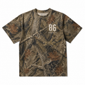A short-sleeve crewneck t-shirt featuring a detailed woodland camouflage pattern with branches, leaves, and bark. The front has a white printed graphic reading '86 SOCIETY'.