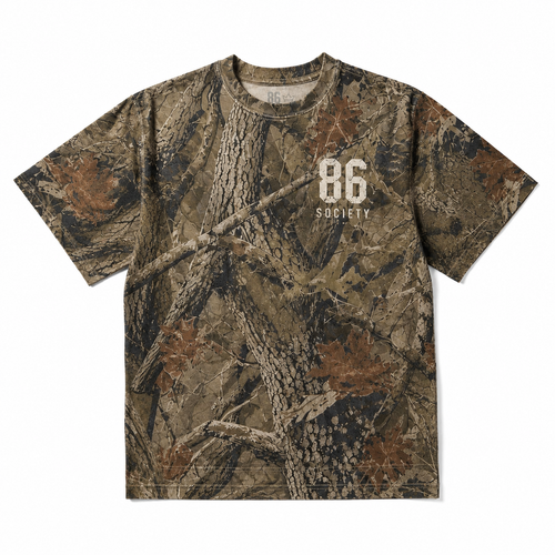 A short-sleeve crewneck t-shirt featuring a detailed woodland camouflage pattern with branches, leaves, and bark. The front has a white printed graphic reading '86 SOCIETY'.